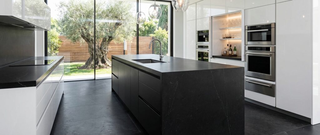 Cocina con isla lujosa de Sabaya inspirada en cocinas con isla, con una gran isla negra con fregadero integrado, electrodomésticos empotrados y un ventanal que conecta con el jardín; una imagen elegante que muestra cómo distribuir cocinas con isla de alta gama e invita a descubrir más proyectos haciendo clic en ella.