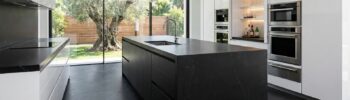 Cocina con isla lujosa de Sabaya inspirada en cocinas con isla, con una gran isla negra con fregadero integrado, electrodomésticos empotrados y un ventanal que conecta con el jardín; una imagen elegante que muestra cómo distribuir cocinas con isla de alta gama e invita a descubrir más proyectos haciendo clic en ella.