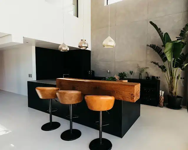 Cocina de isla negra con barra de madera maciza y taburetes de cuero, en un espacio de doble altura