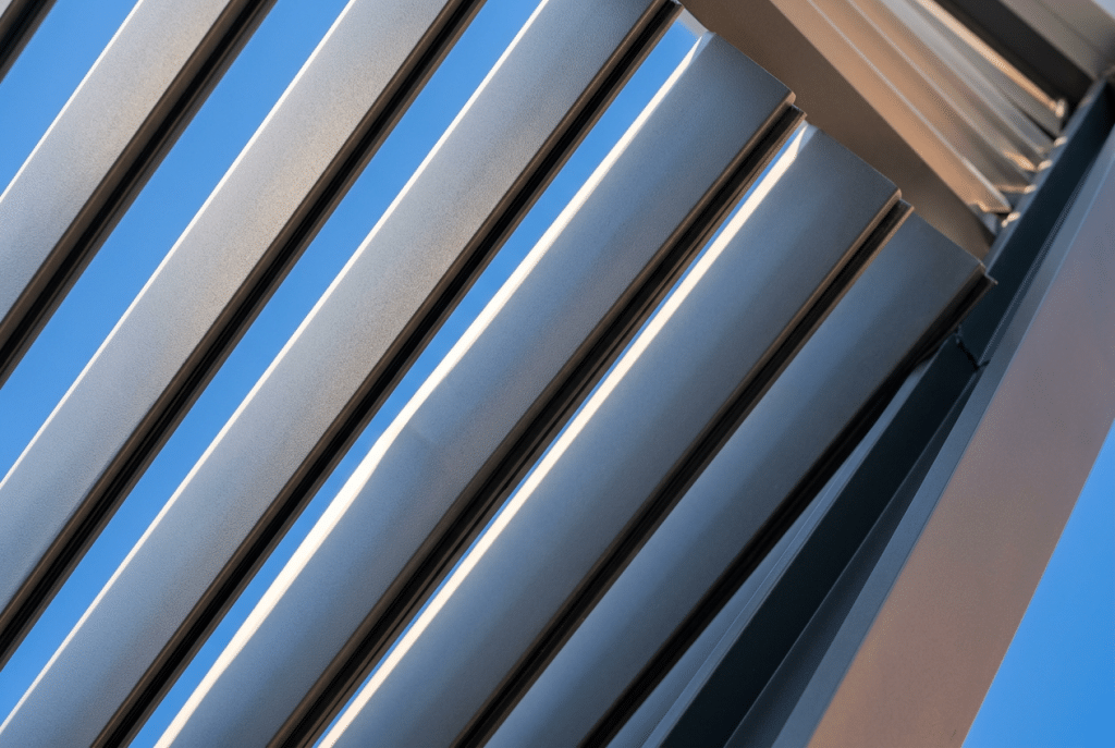 Lamas de aluminio antracita de una pérgola bioclimática vistas desde abajo, giradas a medio abrir, con el sol de mediodía colándose entre ellas